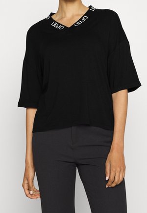 Persona que lleva una blusa negra de manga corta y suelta con un escote en V que presenta el texto repetido "LIU·JO" en blanco alrededor del cuello.