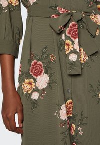 Olivgrünes Kleid mit floralen Mustern in Pink, Rot und Gelb. Verfügt über einen Taillengürtel und eine Knopfverzierung, strukturierter Stoff mit einem leichten Glanz.