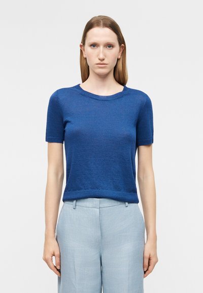 Femme aux cheveux châtains clairs et raides, portant un haut en maille bleu à manches courtes et un pantalon taille haute gris clair, debout devant un fond uni.