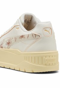 Valkoinen Puma-tenniskenkä beige pohjalla, kukkakuosisella mokkanahka-raidalla ja kohokuvioidulla Puma-logolla kantapäässä ja sivussa.