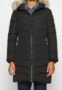 Černý péřový kabát s předním zipem, horizontálním prošívaným designem, odnímatelnou kapucí s kožíškem, bočními kapsami a žebrovanými manžetami.