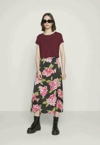 Mulher a usar óculos de sol pretos, uma blusa curta de manga na cor burgundy, uma saia midi preta com estampado floral rosa e botas de combate pretas, de pé.
