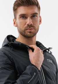 Sort letvægtsjakke med hætte og lynlås, justerbare snore, kontrasterende indvendig for og trykt tekst på brystet.