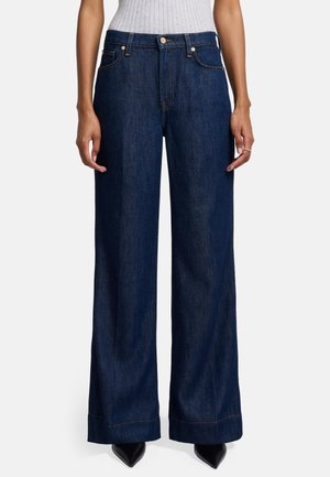 Brede denim jeans in donkerblauw. Kenmerkt een hoge taille, voorzakken en zichtbare stiksels. Gecombineerd met puntige zwarte hakken.