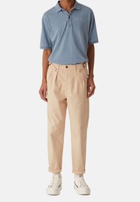 Muž oblečený v pískově modré polo tričko, béžových plisovaných kalhotách s nastavitelnými postranními popruhy, bílých ponožkách a bílých teniskách na bílém pozadí.