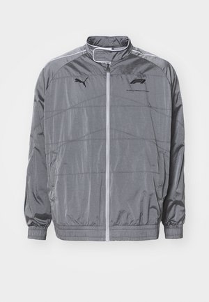 Giacca grigia con zip realizzata in materiale liscio e leggero. Presenta linee di design, polsini elasticizzati e un logo in evidenza sul petto.