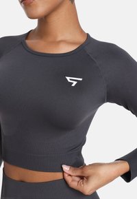 Crna atletska majica dugih rukava s uskim krojem. Ima rebrastu teksturu, okrugli izrez i bijeli logo na lijevoj strani prsnog koša.
