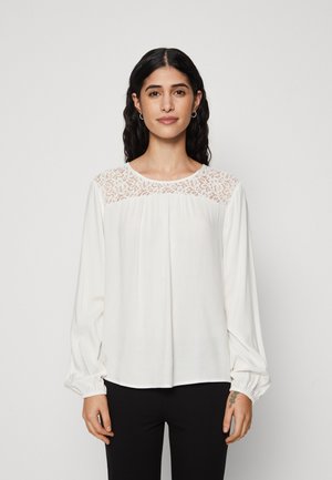White long-sleeve blouse with lace detailing on the shoulders, gathered cuffs, and a relaxed fit, paired with black pants.