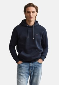 Sweat à capuche bleu marine avec une poche kangourou, des cordons de serrage et un logo sur la poitrine. Confectionné en tissu doux, avec des poignets et un ourlet côtelés.