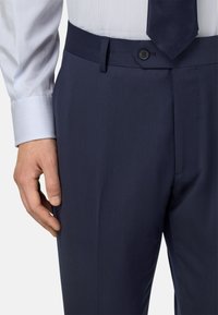 Jūrinės spalvos kelnės, pagamintos iš glotnaus audinio, su plokščiu priekiniu dalimi, diržo kilpomis ir šoniniu kišeniu. Matoma juostuota marškinėlių rankovė.