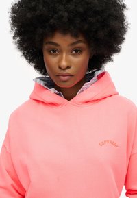 Sweat à capuche rose avec cordon de serrage, arborant le nom de la marque "Superdry" brodé en orange. Superposé sur un col de chemise à carreaux.