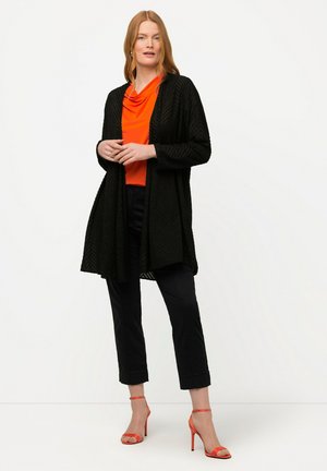 Cardigan largo negro con estampado, superpuesto sobre una blusa naranja de cuello vuelto, combinado con pantalones negros recortados y sandalias de tacón rojas.