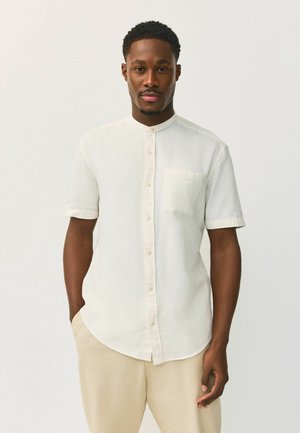 Mann mit kurzen, lockigen Haaren, der ein weißes kurzärmliges Hemd mit Knöpfen und beige Hosen trägt, stehend mit einer Hand in der Hosentasche.