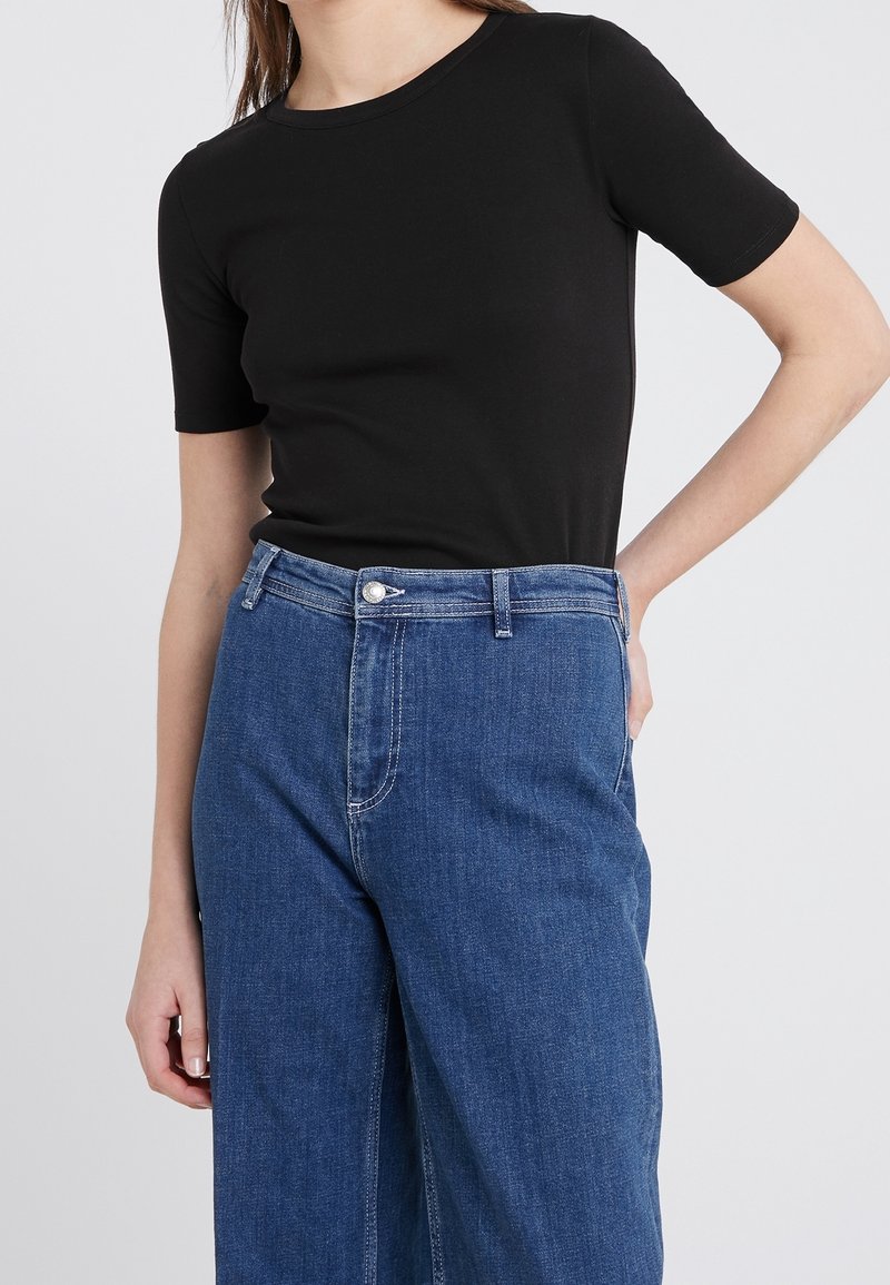 Jeune personne portant un t-shirt noir ajusté à manches courtes, rentré dans un jean bleu large taille haute, debout avec une main sur la hanche.