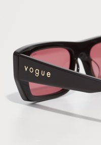 Čierne obdlžnikové slnečné okuliare s ružovými sklami a zlatým logom "vogue" na stranici, zobrazené na bielom pozadí.
