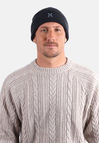Homme portant un bonnet en tricot noir et un pull en tricot beige à motif torsadé, faisant face à l'avant sur un fond clair uni.