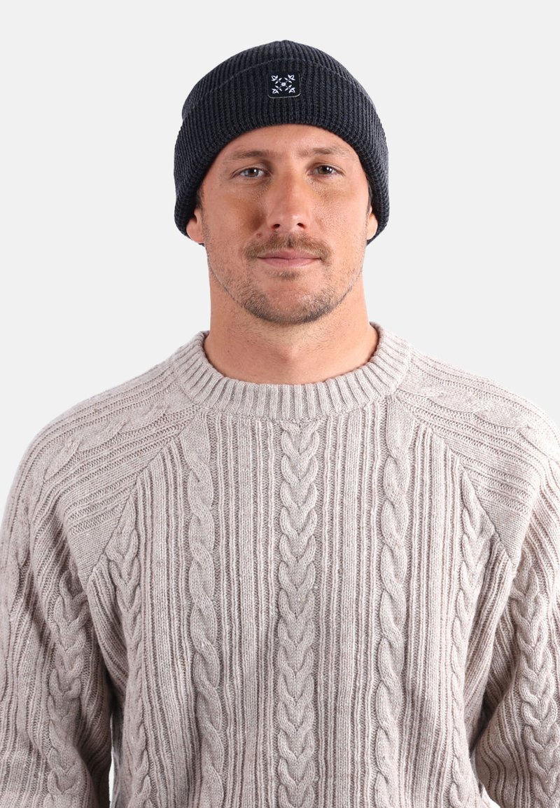 Homme portant un bonnet en tricot noir et un pull en tricot beige à motif torsadé, faisant face à l'avant sur un fond clair uni.