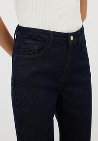 Jeans oscuros de mezclilla con corte ajustado, que cuentan con bolsillos delanteros y traseros y un botón dorado en la cintura.