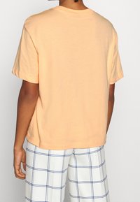 Osoba oblečená v světle broskvovém tričku s krátkým rukávem a bílých kalhotách s modrým kostkovaným vzorem, zobrazená zezadu na jednobarevném pozadí.