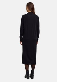 Robe pull noire longue en tissu doux et texturé. Présente une coupe décontractée, des manches longues et un ourlet légèrement arrondi. Portée avec des bottines.