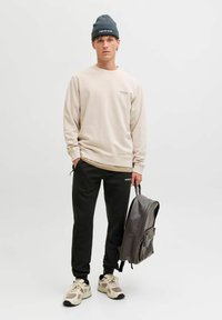 Beige sweatshirt met ribgebreide manchetten, zwarte joggingbroek met zijstrepen, beige en grijze sneakers, en een donkergrijze rugtas.