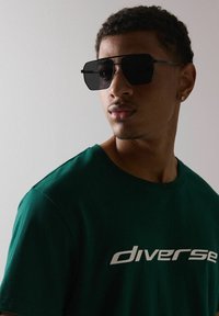 Zielona bawełniana koszulka z białym napisem "diverse", w komplecie z kanciastymi czarnymi okularami przeciwsłonecznymi. Gładka tekstura i luźny krój.