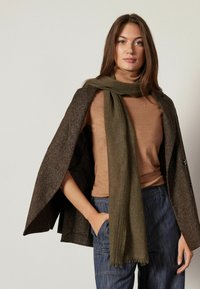 Olivgrön scarf i mjukt tyg, draperad runt halsen. Bärs tillsammans med en figursydd brun topp och en strukturerad brun kappa.