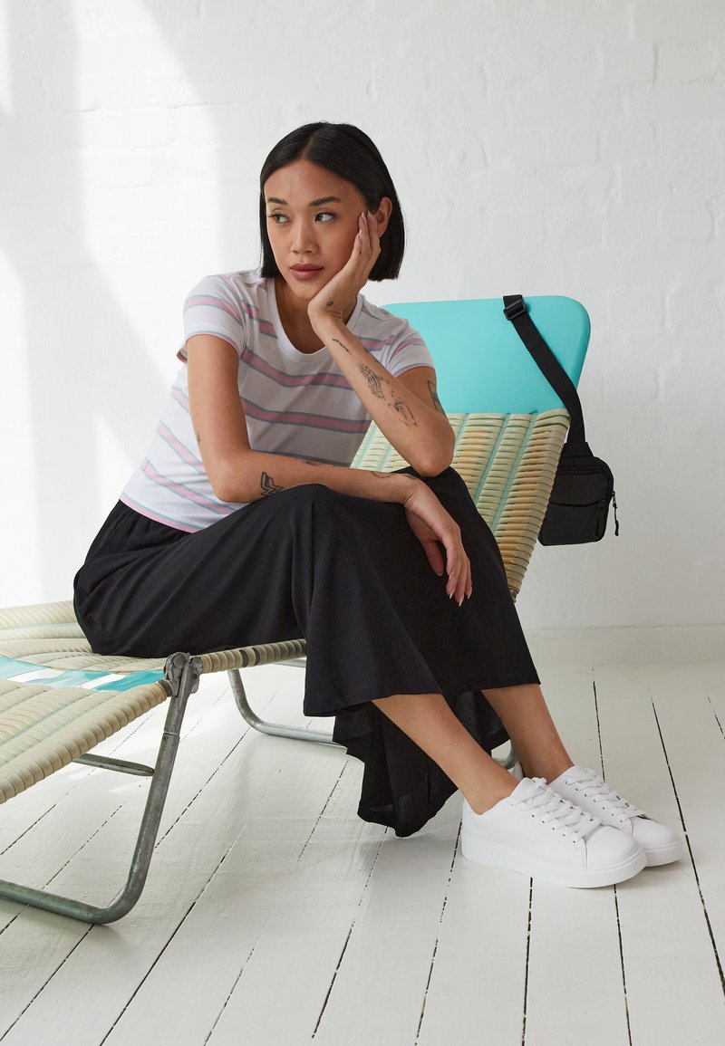Striped short-sleeve shirt in white, pink, and grey; black skirt; white sneakers. Relaxed pose on a woven lounge chair with a teal accent.