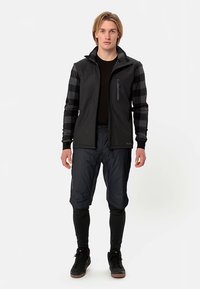 Zwarte softshelljas met een ritssluiting aan de zak, voorzien van zwart-grijs gestreepte mouwen. Gecombineerd met een donkere broek en zwarte schoenen.