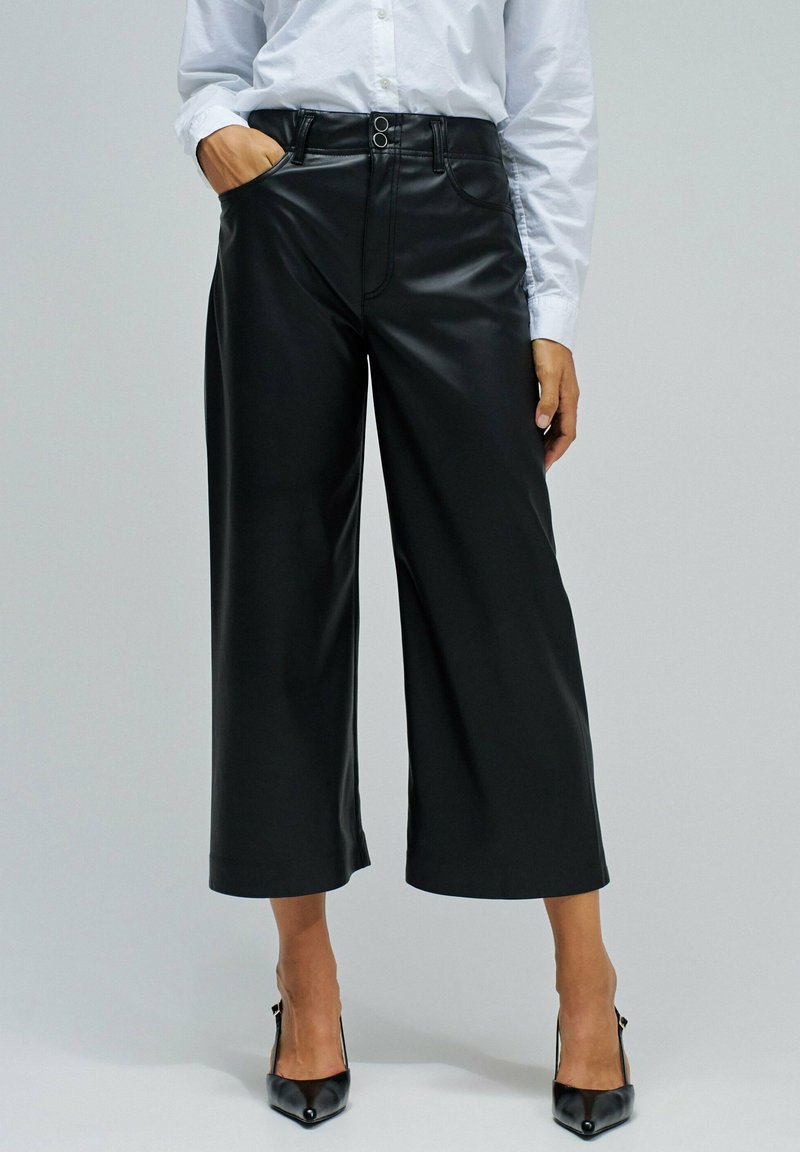 Mujer vestida con pantalones de cuero negros de pierna ancha y cortos, camisa blanca de manga larga metida por dentro y tacones altos negros de punta, de pie con la mano en el bolsillo.