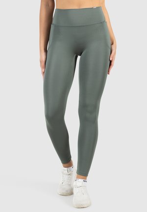 Legginsy z wysokim stanem, prążkowane, w stonowanej zielonej tkaninie, o eleganckim kroju i bocznymi kieszeniami, zestawione z białymi butami sportowymi.