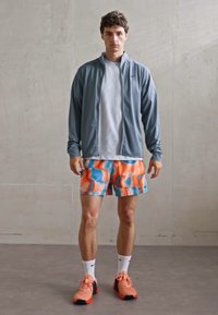 Ropa deportiva que incluye una chaqueta gris, una camiseta blanca, pantalones cortos de patrón vibrante en naranja y azul, calcetines blancos y zapatos en naranja y negro.