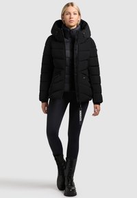 Jachetă puffer neagră cu glugă, cu un design matlasat, buzunare cu fermoar și o siluetă adaptată. Asortată cu colanți negri și botine.