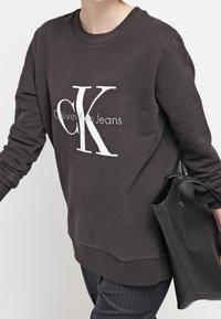 Felpa scura in misto cotone, con un grande logo bianco di Calvin Klein. Maniche lunghe, scollatura rotonda e vestibilità rilassata.