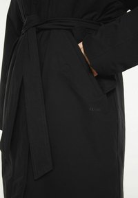 Svart kappa med bälte, som har en ficka och en subtil logotyp. Tillverkad av slät tyg, med en skräddarsydd passform och rena linjer.