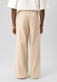 Beige broek met wijde pijpen van een gestructureerde stof, met twee achterzakken en een hoge taille, gezien van achteren.