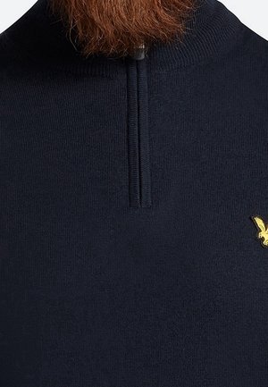 Homme portant un pull en maille bleu marine avec une fermeture éclair courte et un petit logo jaune brodé sur la poitrine.