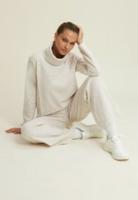 Maglietta a maniche lunghe di un beige chiaro e texture, con colletto alto, abbinata a pantaloni a gamba larga dello stesso colore e scarpe da ginnastica bianche. Posizione seduta.