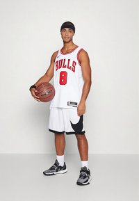 Valkoinen koripallopaita, jossa punainen "BULLS"-teksti ja numero 8, yhdistetty mustavalkoisiin shortseihin. Pitelee ruskeaa Spalding-koripalloa.