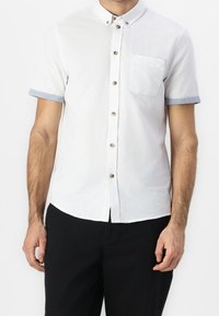 Camicia bianca a maniche corte con bottoni, con polsini blu, taschino sul petto e bottoni marroni. Il materiale sembra leggero e ha una finitura strutturata.