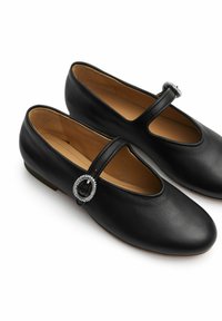 Zapatos Mary Jane de cuero negro con un acento de hebilla redonda, punta redondeada, suela plana y textura suave, presentando un diseño de correa lateral.