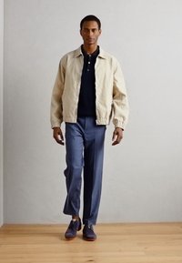 Giacca bomber beige chiaro sopra una polo blu navy, abbinata a pantaloni blu su misura e scarpe scure, in un ambiente minimalista.