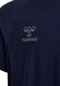 Tissu de chemise de sport bleu foncé montrant des coutures surpiquées et un logo hummel argenté réfléchissant sur le côté gauche de la poitrine.