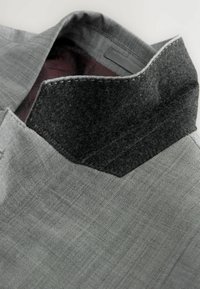 Chaqueta gris con una tela texturizada y un cuello de fieltro negro en contraste. El interior presenta un forro burdeos con un patrón.