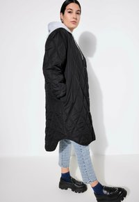 Cappotto lungo trapuntato nero con cappuccio, chiusura a bottoni, tasche laterali, abbinato a jeans in denim blu e scarpe platform nere chunk con borchie.