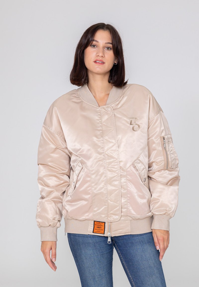 Satinowa bomberka w jasnym beżu, z zapięciem na zamek, bocznymi kieszeniami, ściągaczami przy mankietach oraz pomarańczową metką z przodu.