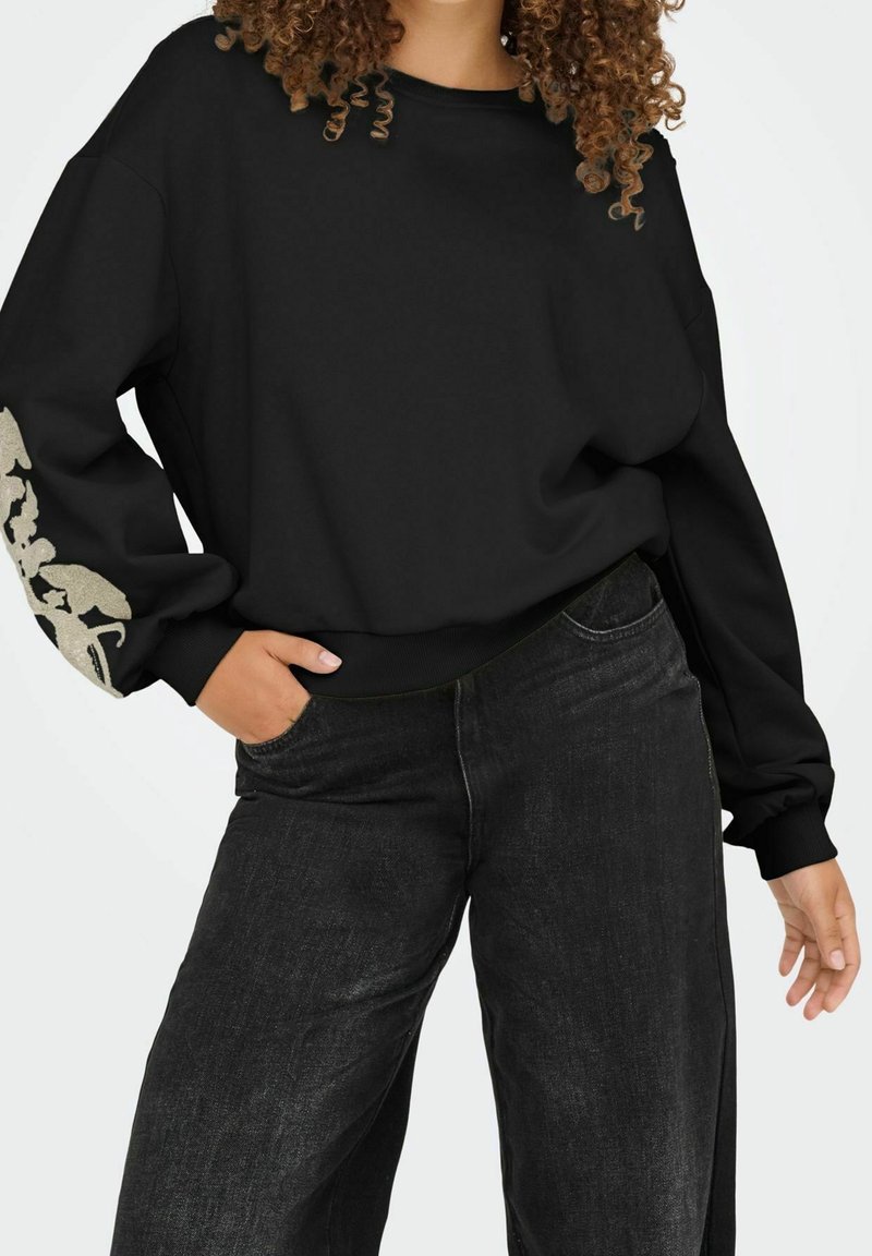 Personne portant un sweatshirt noir ample avec un motif floral beige sur une manche et un jean noir à jambes larges, posant avec une main dans une poche.