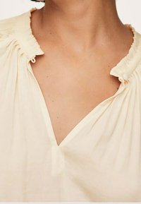 Blusa color crema con scollo a V, con dettagli arricciati attorno al collo e in tessuto morbido e leggero.