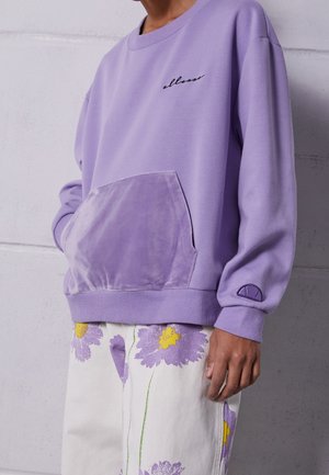 Persona che indossa una felpa viola chiaro con una tasca frontale in velluto e pantaloni bianchi a fiori grandi viola e gialli.