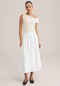 Svijetla kremasta majica s jednim ramenom u kombinaciji s lepršavom, bijelom suknjom do gležnjeva. Odjeća se odlikuje mekim materijalom i naboranim pojasom. Smeđe ravne cipele.